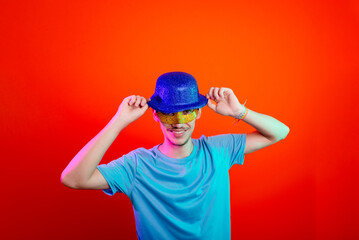 Happy and fun young man in blue shirt, wearing carnival mask and holding blue hat. Posing for the camera. Isolated on neon orange background.