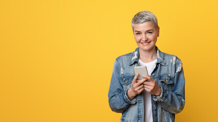 Portrait of beautiful smiling middle aged woman with smartphone in hands over yellow studio background, copy space