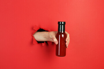 Woman holding bottle of ketchup through hole in torn red paper, closeup