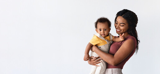 Motherhood Concept. Portrait Of Smiling Young Mother Holding Adorable Infant Son On Arms, Happy African Mom Enjoying Time With Her Baby, Standing Over White Background, Panorama With Copy Space