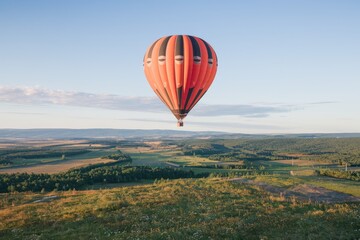 Naklejka premium Orange/Black hot air balloon ascends above a lush, green, rural landscape with soft blue sky. Peaceful, tranquil, travel, adventure