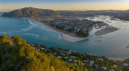 Coastal Pauanui nestled beside a bay, with homes lining the shore and a sandy beach. Aerial view of a tranquil seaside community.  Tairua, Coromandel Peninsula, New Zealand