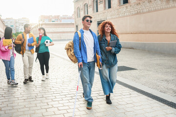 Happy blind man walks with group of friends inside school or university campus. People students,...