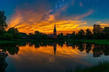 Serene sunset over a picturesque church reflected in a tranquil lake, Milky Way visible in the night sky.