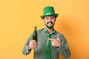Young man in leprechaun hat with green beard pointing at bottle of beer on yellow background. St....