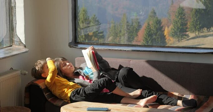 Little kid and mother lying on sofa reading a book in a mountain rustic chalet, enjoy a good time together