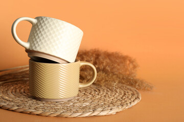 Different ceramic mugs with dry common reeds and wicker mat on orange background
