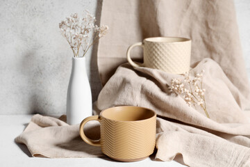 Different ceramic mugs with dried flowers on light grunge background