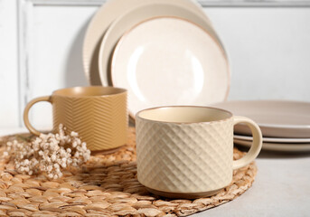 Different ceramic mugs with dried flowers and wicker mat on light background