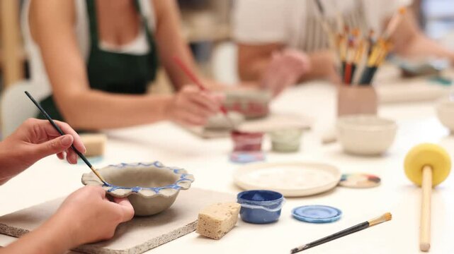 Girl paints bowl of clay during master class, top view. Unrecognizable people team of creative enthusiasts is engaged in clay modeling lesson. Employee covers gray, damp clay bowl with paint
