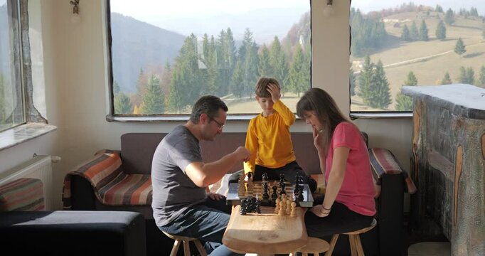 Parents play chess in a mountain rustic chalet, son arbitrates the game