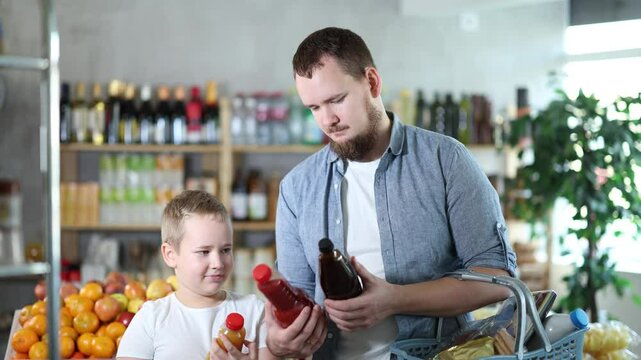 Father with boy hold packages plastic bottle and choose consider fruit juice in store. Market visitors choose make choice and view bottle juice. Client hesitates before buying, chooses drink
