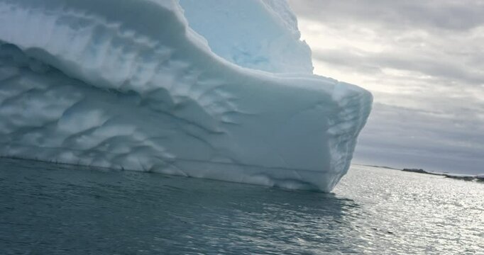 A close-up view of a towering iceberg captured from a boat in Antarctica. The iceberg&rsquo;s intricate textured surface contrasts with the calm waters and cloudy sky.