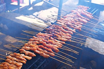 A close-up of rows of chicken satay being grilled on a traditional charcoal barbecue. The skewers...