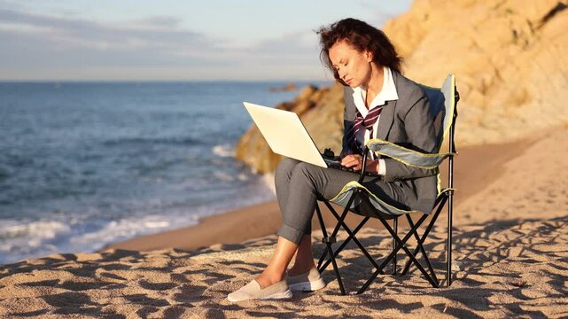Businesswoman in office clothes working on laptop on seashore in sunlight. High quality 4k footage
