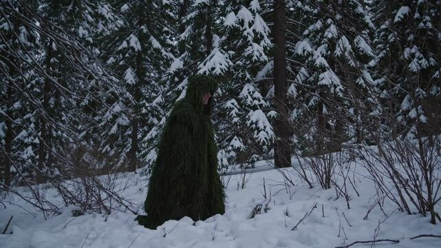 A person in a ghillie suit blends into a snowy forest. The low-angle shot captures the serene, camouflaged scene, ideal for a nature-themed video.