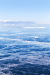 Obraz premium Typical winter landscape of frozen Baikal Lake. The endless fields of smooth blue ice with cracks and snowy mountains in the distance. Natural background. Winter travels and outdoors