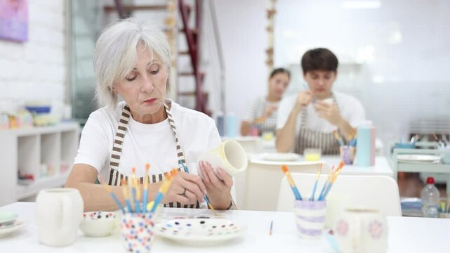 Interested elderly woman attending pottery class, enthusiastically painting on ceramic cup, creating unique custom design. Creative hobby concept