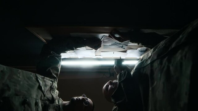 Low key view from below of two commanders in camouflage uniform discussing military plan while standing by board with maps and documents
