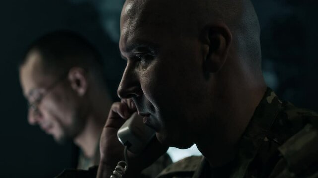 Low key close up shot of military officer talking on landline phone while his colleague analyzing data in command center