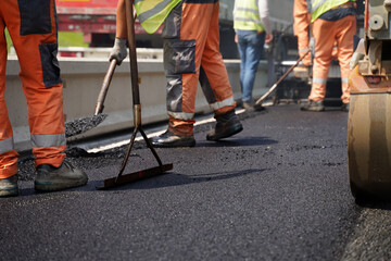 Low-angle shot of multiple construction workers’ legs using tools to spread fresh asphalt next to a road barrier, highlighting teamwork and precision in road construction near barriers. © Redline Visuals