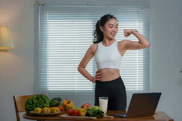 Happy athletic woman flexing her biceps while following an online workout session on a laptop and...
