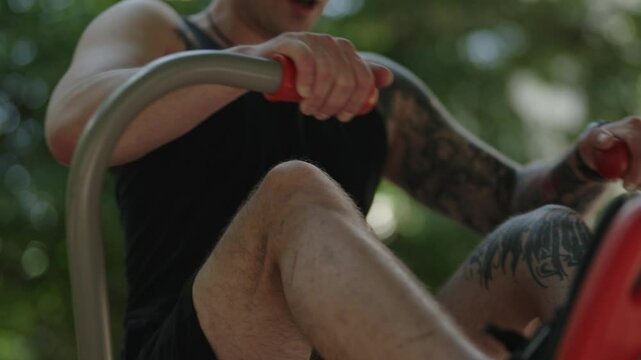 A man working out on a rowing machine at an outdoor fitness park. He is wearing a black tank top and shorts, and has tattoos on his arms.