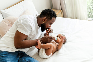 Loving African father bottle-feeding his baby in a cozy bedroom. Tender moment of bonding, care, and fatherhood. Parenthood, baby care, and family love concept in a warm, nurturing home setting.