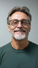 Obraz premium close-up of a smiling, mature man with gray hair, glasses, and a beard, wearing a green shirt against a neutral background.