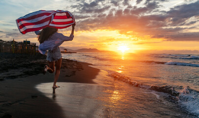 Beautiful patriotic young woman with the American flag held in her standing in the summer sunset