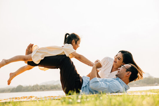 Family bonds over laughter and joy in the park. Father lifts his daughter high, as she happily flies like a plane with arms spread wide, and mother watches with pride. A picture of family fun and love