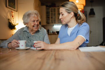 Lovely eldery woman serving tea to friendly caregiver during home visit.