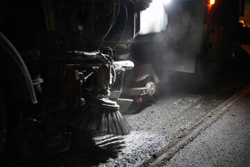 Detailed view of equipment sweeping away dirt on a road construction site at night, after the old asphalt was milled away, preparing the surface for the next phase of roadwork.