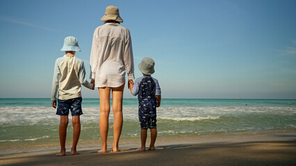 A mother and her two children stand on the shore, holding hands while gazing at the ocean waves. The sky is clear, and the sun casts a warm glow on the sandy beach.