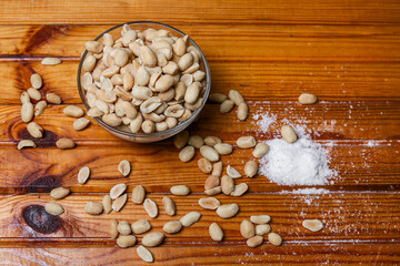Close-up of salted peanuts in a bowl on a wooden board. Salt grains and nuts are scattered on the surface. View from above