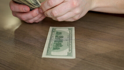 A person is counting crisp dollar bills on a smooth wooden table, surrounded by a calm atmosphere in the afternoon light. The focus is on the careful handling of money.