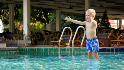 A young boy with short blonde hair is standing in a swimming pool, arms wide open in excitement. The pool is situated in a vibrant resort setting surrounded by lounge chairs.