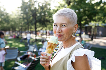Side view outdoor portrait of smiling senior female with short stylish haircut eating tasty...