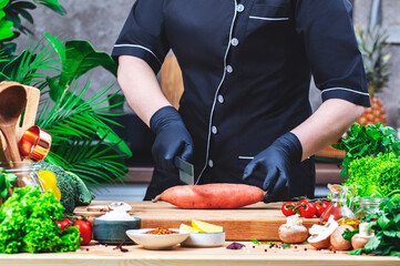 Chef cuts sweet potato for baking. Cozy kitchen with wooden table, kitchenware, vegetables, herbs and ingredients for cooking. Healthy vegan eating, culinary, recipes, food blogging comcept