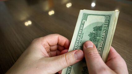 A person carefully counts a stack of hundred dollar bills on a polished wooden table. Soft, natural light highlights the texture of the currency and the person's hands.