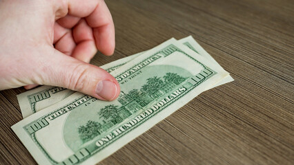 A hand holds several one hundred dollar bills, placing them carefully on a smooth wooden table. This moment captures the act of managing cash, indicating a transaction.