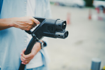Close up of man holds electrical car cable charging with blur car parking at gas station. Eco, renewal energy concept.