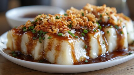 A plate of steamed rice rolls topped with crispy onions and sauce, served with a side dip.