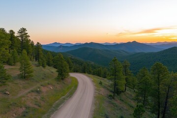 Naklejka premium A road winds through a forested area with a beautiful sunset in the background. The sky is a mix of orange and pink hues, creating a serene and peaceful atmosphere. The trees lining the road are tall