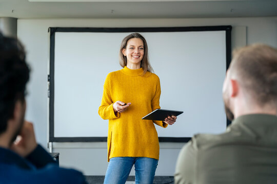 Smiling businesswoman standing with tablet PC and giving presentation to colleagues in office