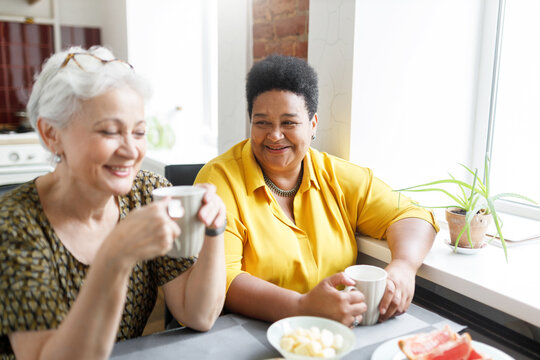 Two senior besties of diverse ethnicity drinking coffee together at kitchen table, caucasian one laughing at her african american best friend's joke. Elderly females having breakfast at kitchen