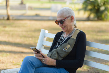 Fototapeta premium Middle-aged lesbian woman smiles sitting on a park bench while checking her cell phone