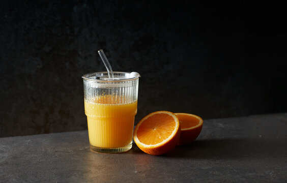 Glass of orange juice with glass straw and halved oranges on dark background