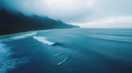 Aerial view of surfers riding waves near a misty mountain coastline