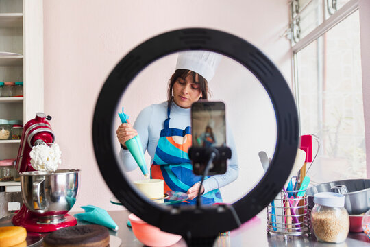 Young confectioner decorating cake and filming tutorial video through smart phone at home